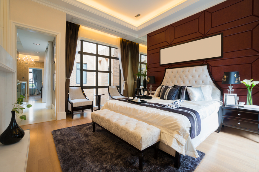 Luxurious master bedroom with wood paneled wall, all-white bed, light wood floor and stylish sitting area